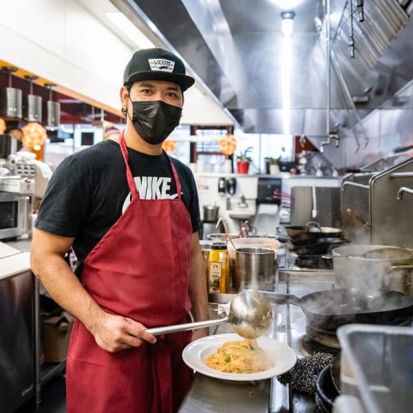 Chef in a black mask and red apron serving pasta from a wok in a busy kitchen.