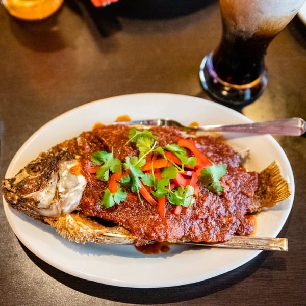 Whole fried fish topped with red sauce, cilantro, and bell peppers on a white plate.