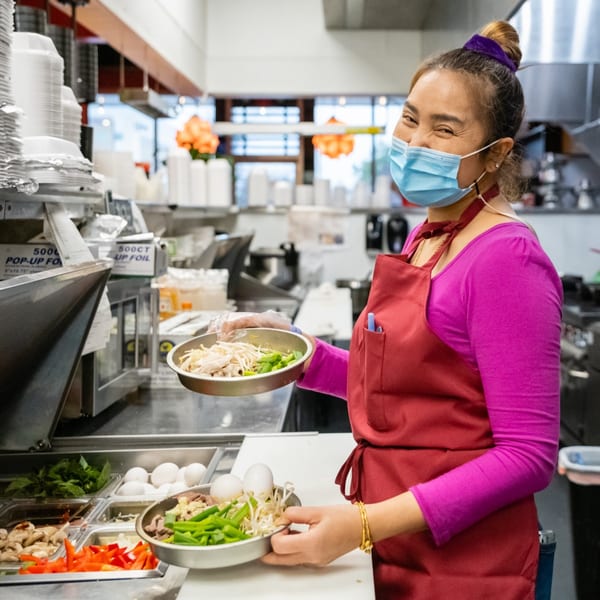 Smiling chef in a face mask and red apron holding two bowls of fresh ingredients in a kitchen.