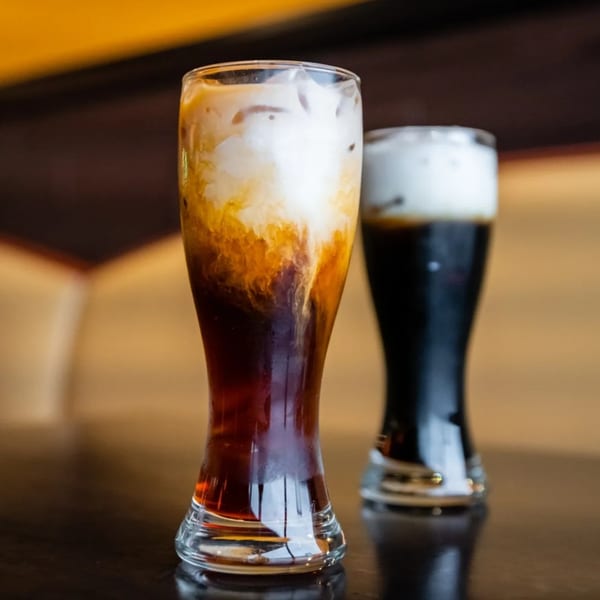 Two tall glasses of iced coffee, one with cream swirling into dark coffee, on a dark table.