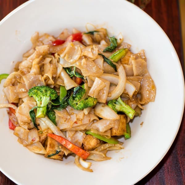 Close-up of a white bowl filled with Pad See Ew, featuring wide noodles, broccoli, and onions.
