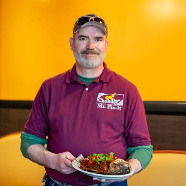 A man in a "Chokdee Mr. Fix-It" shirt holds a plate with a whole fried fish.