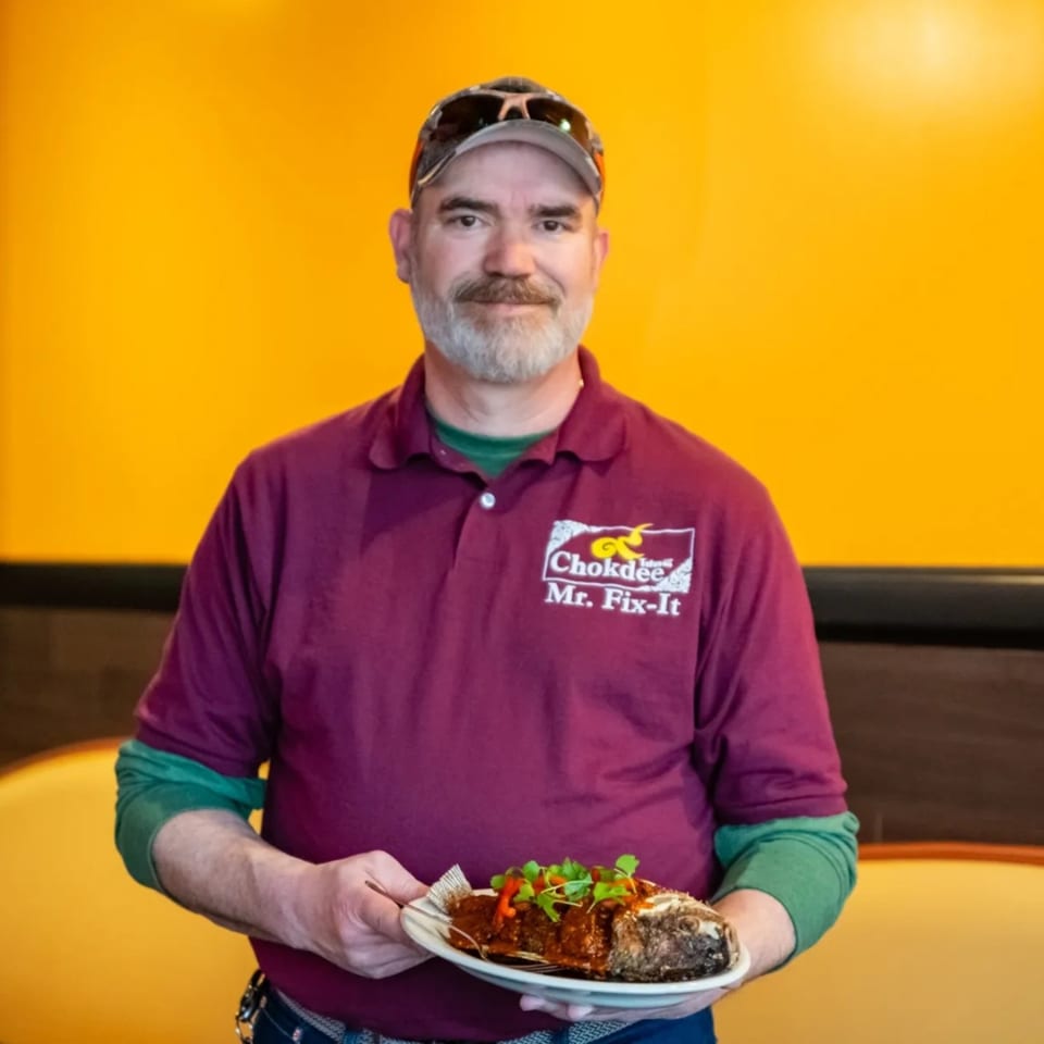A man in a "Chokdee Mr. Fix-It" shirt holds a plate with a whole fried fish.