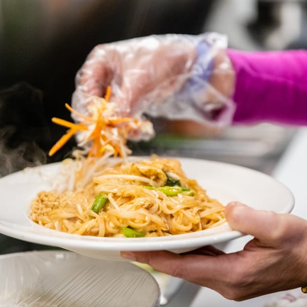 Gloved hands garnishing a steaming plate of Pad Thai noodles with shredded carrots and cabbage.