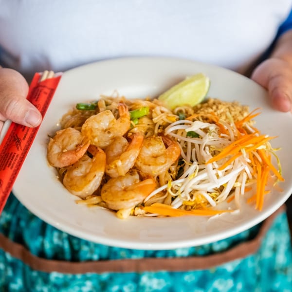 Hands holding a white plate of shrimp pad Thai with bean sprouts, carrots, and a lime wedge.