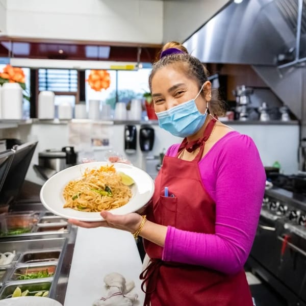 Smiling chef in a mask and red apron holds a plate of Pad Thai in a restaurant kitchen.