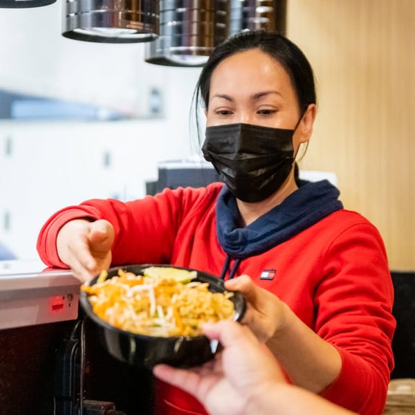 A masked woman in a red hoodie hands a bowl of food to another person.